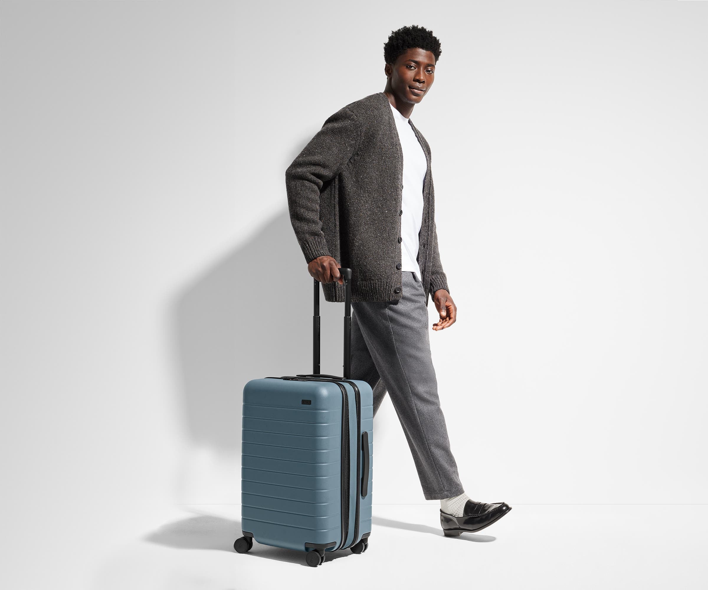 A 6'1" male model is standing next to and walking with The Bigger Carry-On suitcase by holding the raised handle to show size and scale